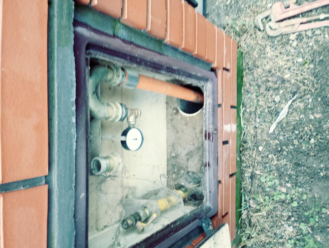 Studzienka rewizyjna z rurami, manometrem i zaworami, otoczona ceglaną obudową, widoczne narzędzia hydrauliczne na ziemi.