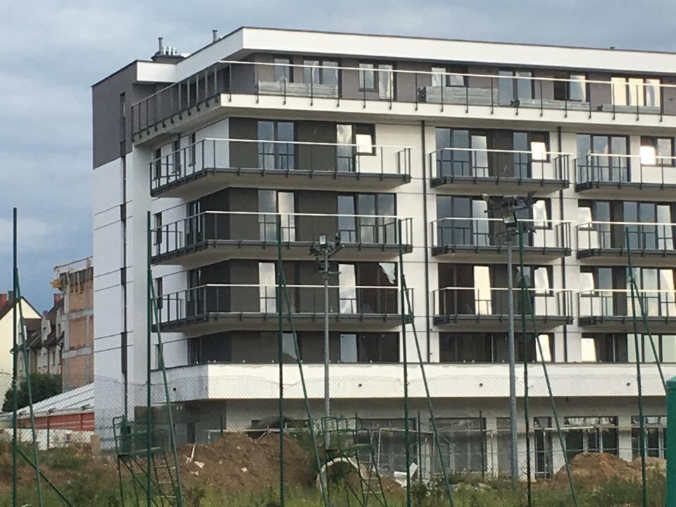 Nowoczesny, czteropiętrowy dom murowany z balkonami w Górnie, widziany zza ogrodzenia budowy i hałdy ziemi.