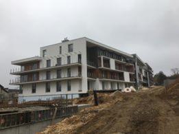 Wielopiętrowy budynek mieszkalny w trakcie budowy, z widoczną elewacją w kolorze białym, drewniane elementy balkonów, ziemia i szalunki w tle.