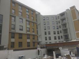 Wielopiętrowy budynek w trakcie prac elewacyjnych, widoczne rusztowania i materiały budowlane, część fasady w kolorze żółtym i białym, pochmurne niebo w tle.