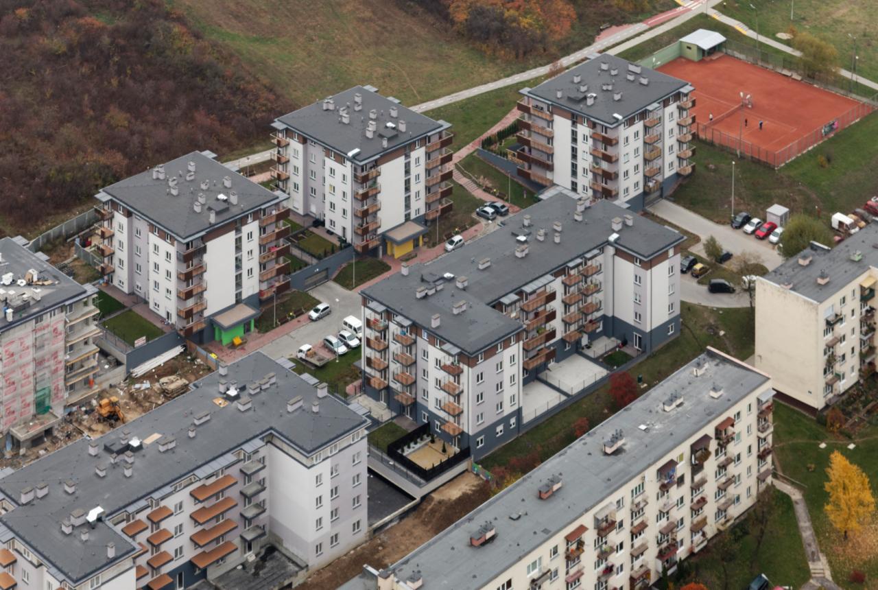 Widok z góry na kompleks nowych, wielopiętrowych budynków mieszkalnych z balkonami, otoczonych zielenią i parkingami, z widocznym kortem tenisowym w tle.