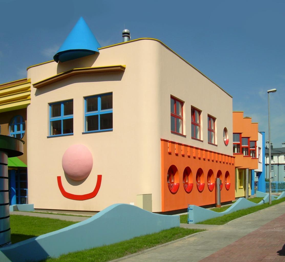 Kolorowy budynek przedszkola z elementami elewacji przypominającymi twarz klauna: niebieski stożek na dachu, okna jako oczy, różowy okrągły nos i czerwony uśmiech. Pozostałe budynki w rzędzie...