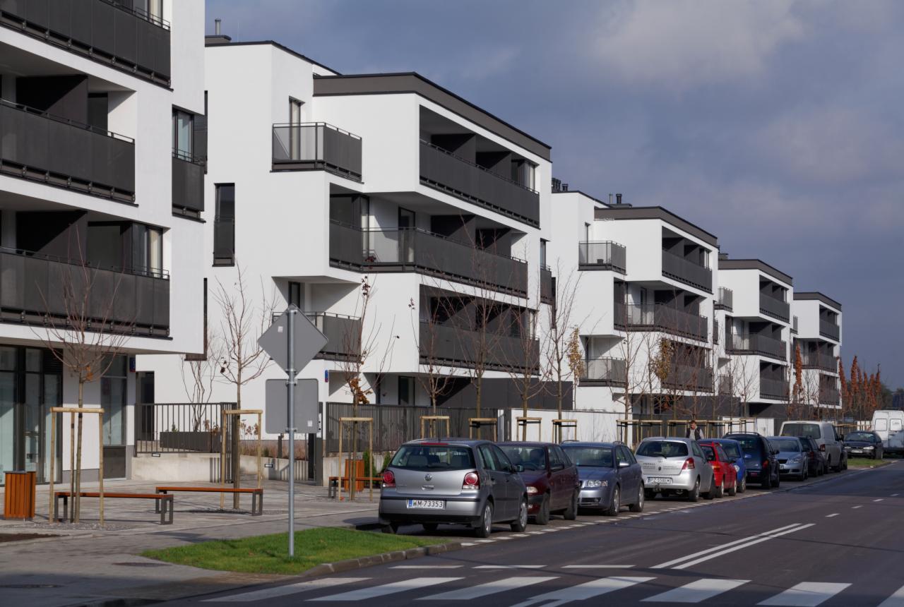 Nowoczesne, białe budynki wielorodzinne z czarnymi balkonami i płaskimi dachami, widok z poziomu ulicy z zaparkowanymi samochodami i przejściem dla pieszych.