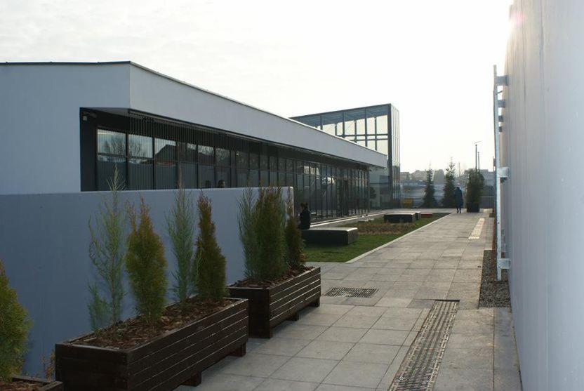 Nowoczesny budynek biurowy z przeszkloną fasadą i jasną elewacją, widok z zewnątrz na chodnik z donicami z iglakami.