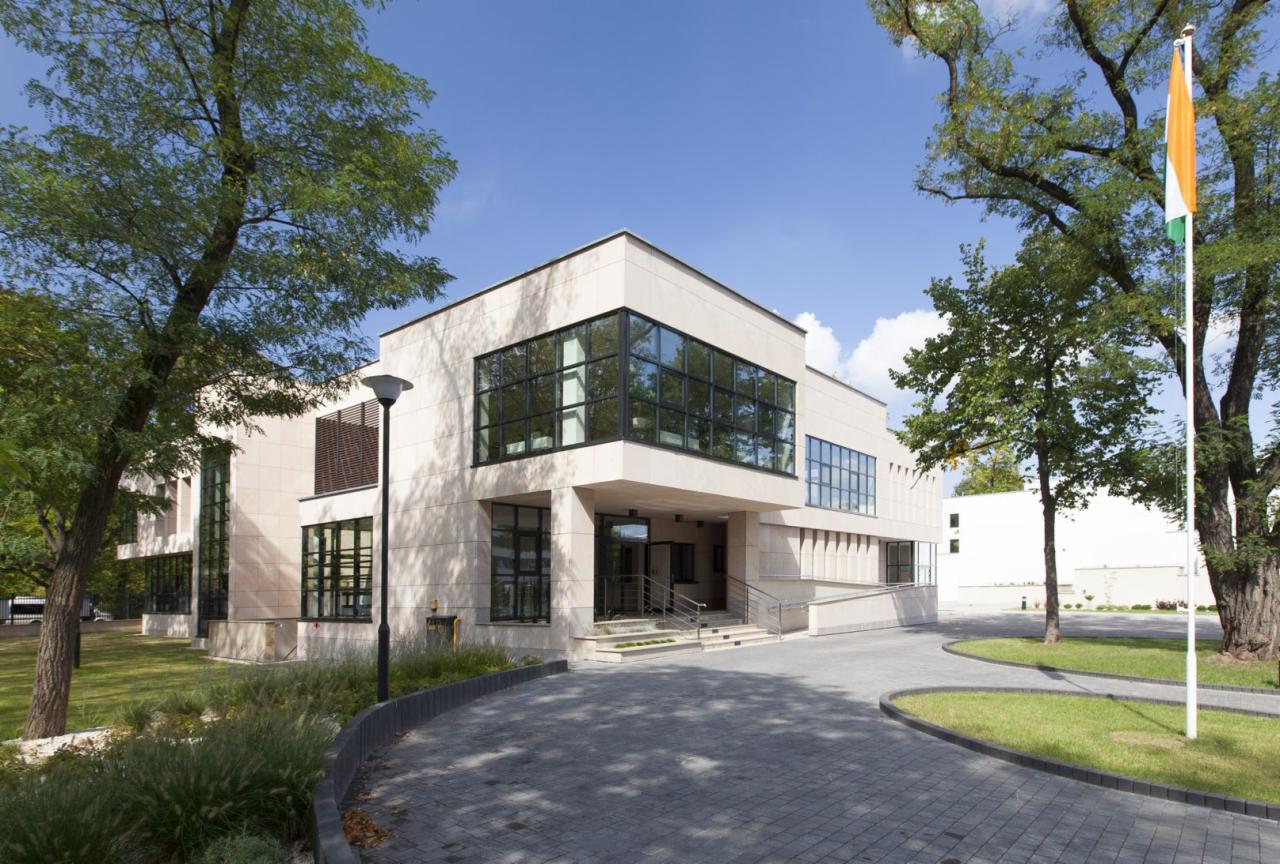 Nowoczesny budynek biurowy z elewacją z jasnego kamienia, duże okna z czarnymi ramami, otoczony zielenią i brukowanym podjazdem.