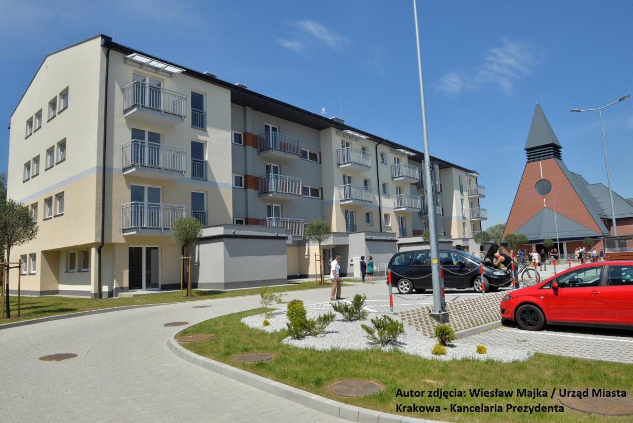 Nowoczesny, trzypiętrowy budynek mieszkalny z balkonami i dwukolorową elewacją, na pierwszym planie zaparkowane samochody i kościół w tle.
