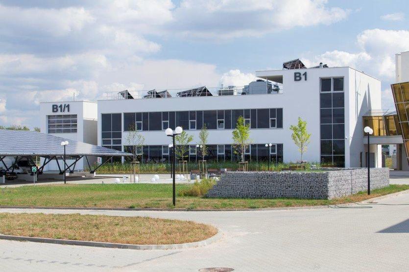 Nowoczesny budynek biurowy z białą elewacją, dużymi oknami, oznaczeniami B1 i B1/1, panele słoneczne na dachu i w wiacie parkingowej, otoczony zielenią i kamiennymi gabionami.