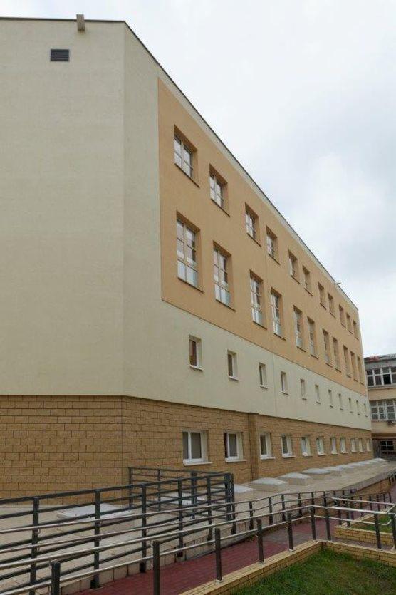 Wielopiętrowy budynek z odnowioną elewacją w odcieniach beżu i brązu, widoczny fragment podjazdu dla niepełnosprawnych i sąsiedni budynek.