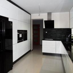 Meble na wymiar - Nowoczesna kuchnia z białymi frontami szafek, czarnym blatem i okładziną z kamienia naturalnego, widoczna czarna lodówka z dystrybutorem wody.