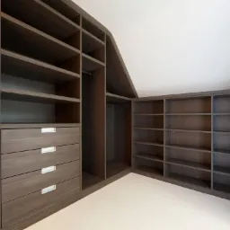 Ciemna, drewniana garderoba z szufladami i półkami, dopasowana do skosu dachu, z minimalistycznymi, srebrnymi uchwytami.