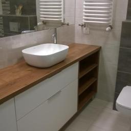 Meble na wymiar - Biała szafka łazienkowa z drewnianym blatem i owalną umywalką nablatową, widoczne otwarte półki z boku, w tle grzejnik drabinkowy.