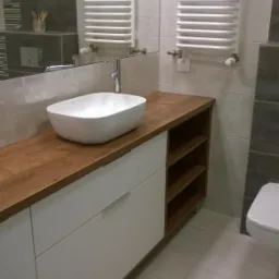 Biała szafka łazienkowa z drewnianym blatem i owalną umywalką nablatową, widoczne otwarte półki z boku, w tle grzejnik drabinkowy.