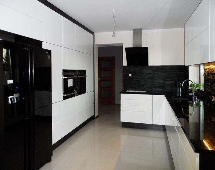 Nowoczesna kuchnia z białymi frontami szafek, czarnym blatem i okładziną z kamienia naturalnego, widoczna czarna lodówka z dystrybutorem wody.