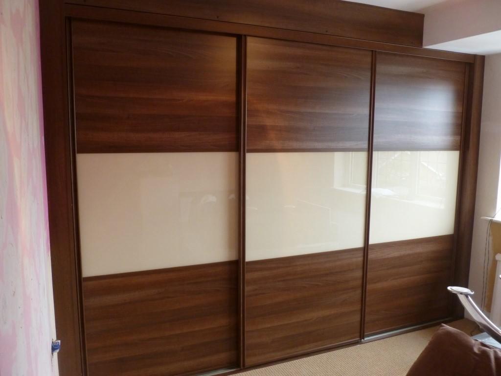 Trzydrzwiowa szafa wnękowa z przesuwnymi drzwiami, fornir w odcieniach brązu i kremowe panele szklane, widoczne odbicie okna w panelach.