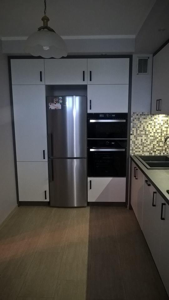 Nowoczesna kuchnia z białymi szafkami, zabudowanym sprzętem AGD (lodówka i piekarnik) oraz mozaiką nad blatem roboczym, oświetlona wiszącą lampą.