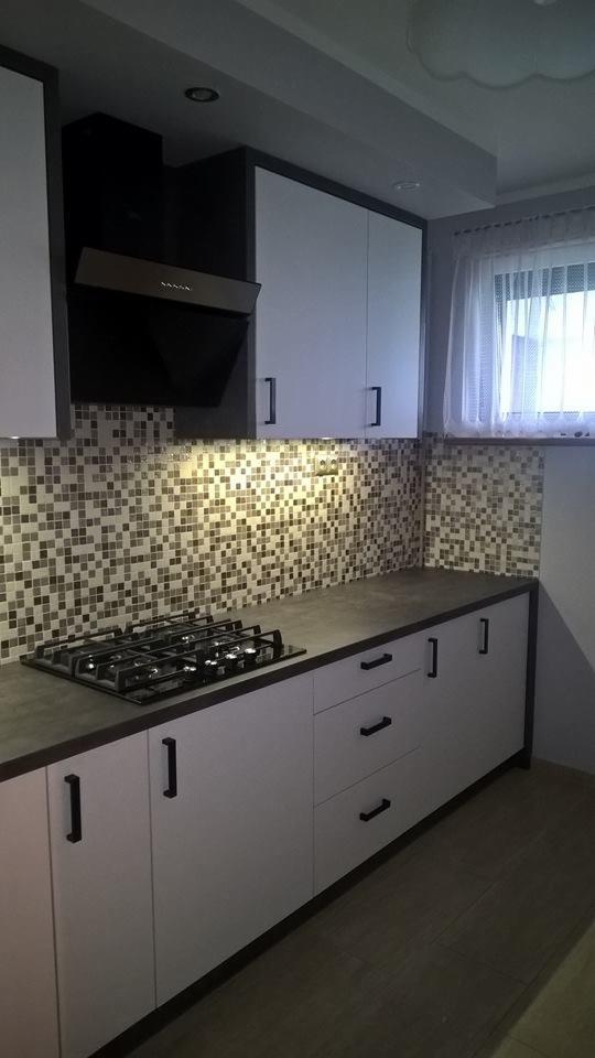 Nowoczesna kuchnia z białymi szafkami, czarnymi uchwytami i mozaikową ścianą nad blatem, oświetlona punktowym światłem, z widoczną płytą gazową.