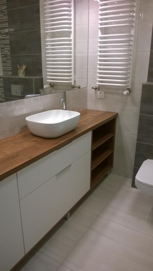 Biała szafka łazienkowa z drewnianym blatem i owalną umywalką nablatową, widoczne otwarte półki z boku, w tle grzejnik drabinkowy.