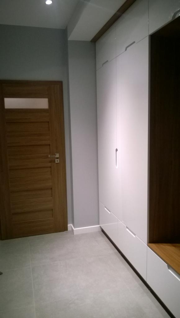 Biała szafa wnękowa z drewnianymi akcentami, minimalistyczne uchwyty, szara ściana i drewniane drzwi z przeszkleniem, szara podłoga z płytek.