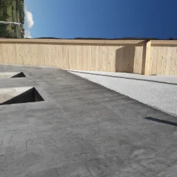 Surowy dach z betonu z otworami, drewniana konstrukcja pionowa w tle, widok z góry, jasne niebo.