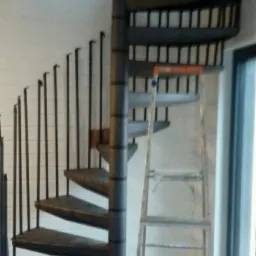 Metalowe schody spiralne w trakcie montażu, widoczna drabina i kartonowe pudełko na podłodze.