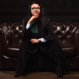 Portret kobiety w todze prawniczej siedzącej na skórzanej sofie, z ręką pod brodą, w okularach i z zegarkiem na ręku, na ciemnym tle.