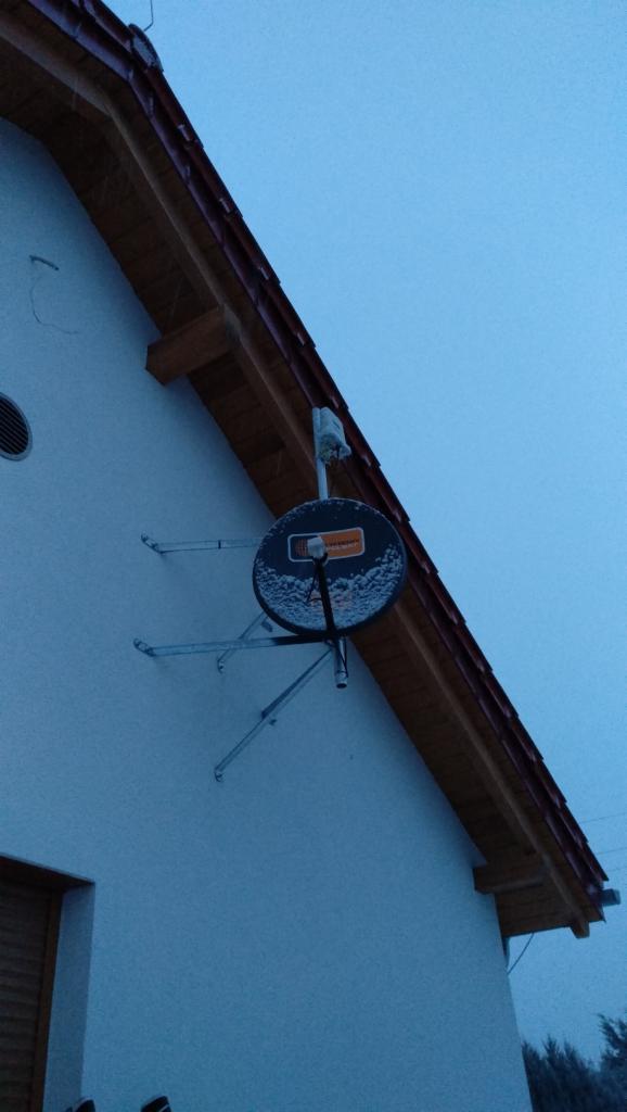 Zewnętrzna antena satelitarna zamontowana na ścianie domu, lekko przyprószona śniegiem, z widocznym kablem antenowym i uchwytami mocującymi.