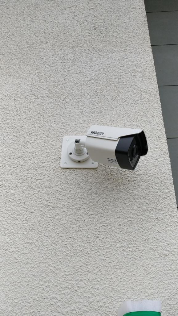 Kamera monitoringu CCTV marki HQ CCTV zamontowana na szorstkiej, jasnej elewacji budynku, widoczna z boku.