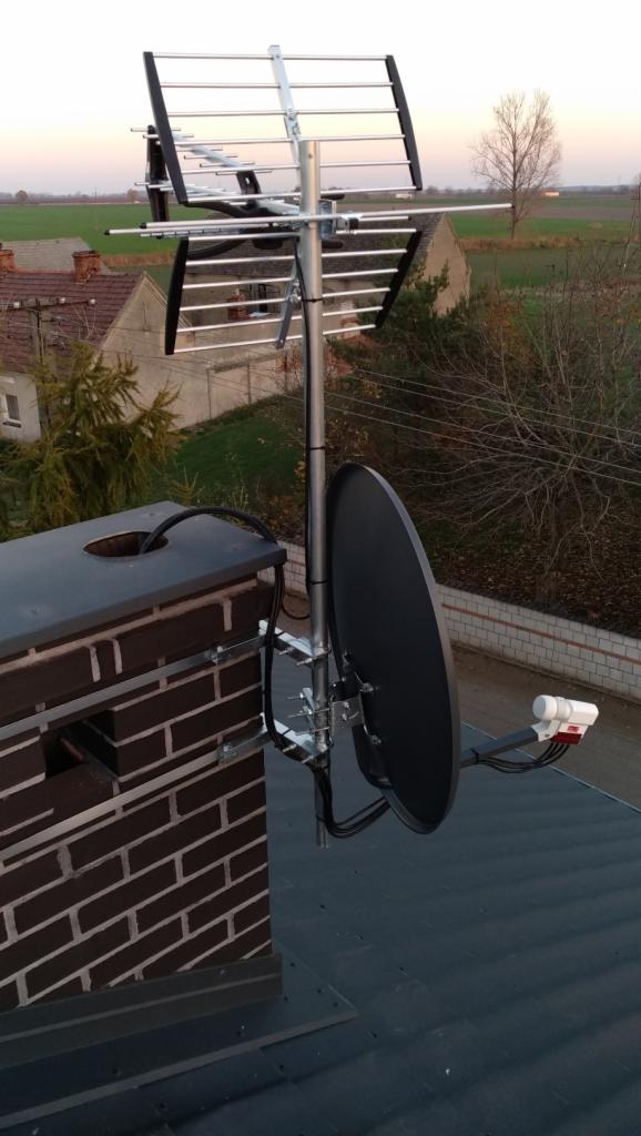 Antena satelitarna i antena kierunkowa zamontowane na kominie z cegły na dachu budynku, widok na okoliczne domy i pola w tle.