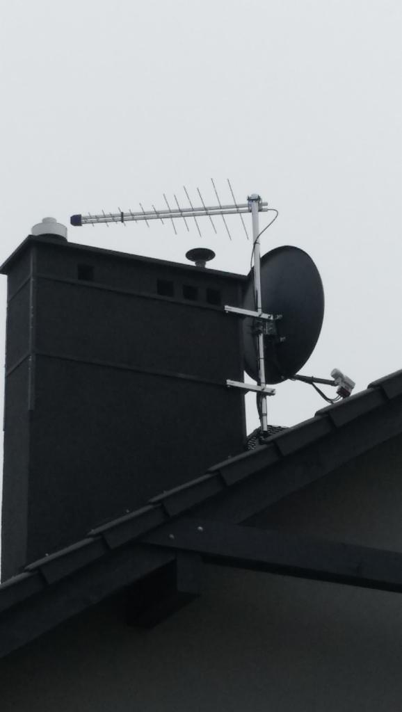 Antena Yagi-Uda i antena satelitarna zamontowane obok komina na dachu domu jednorodzinnego, widok z dołu w pochmurny dzień.