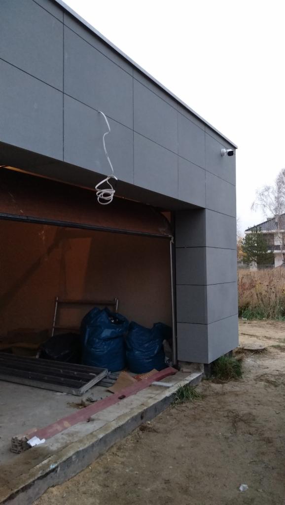 Szary budynek garażu z widoczną kamerą monitoringu na ścianie, otwartą bramą, workami w środku i przewodami wiszącymi z sufitu.