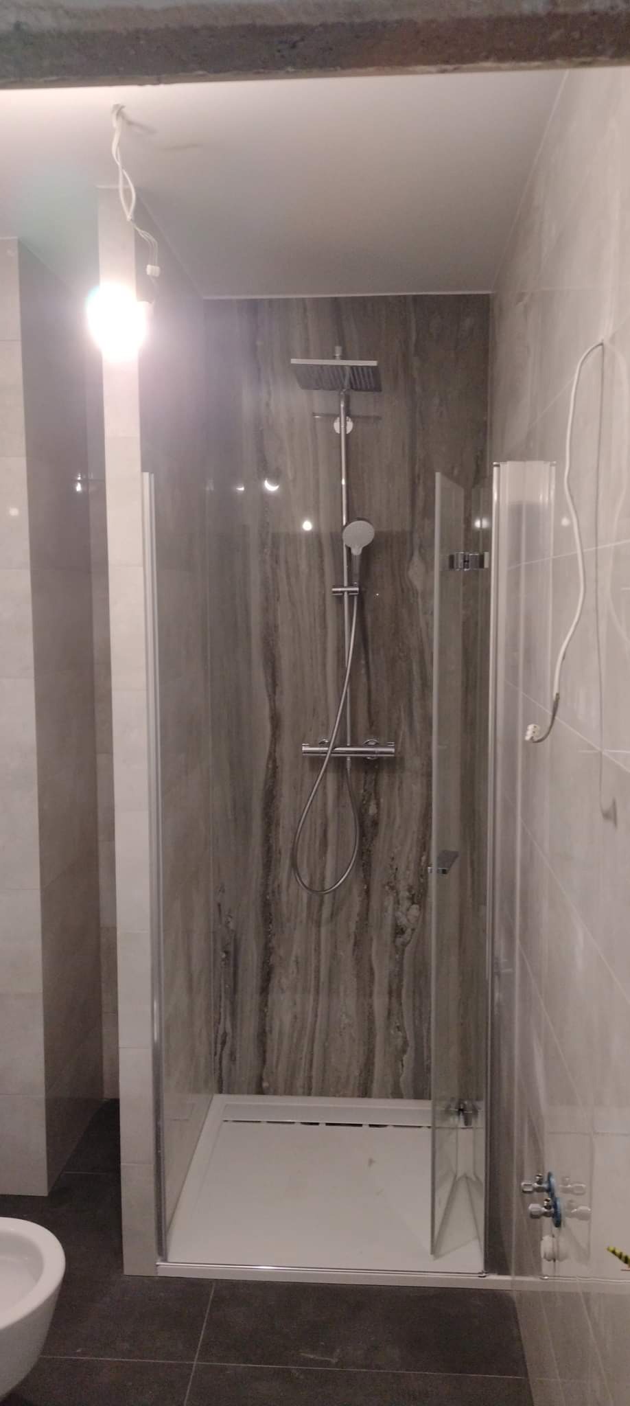 Nowoczesna kabina prysznicowa z deszczownicą i szklanymi drzwiami, wykończona płytkami imitującymi marmur, z widocznymi przewodami elektrycznymi zwisającymi z sufitu.