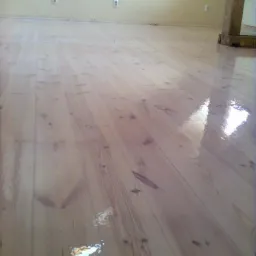 Błyszcząca, świeżo polakierowana podłoga z jasnego drewna, widoczne odbicia światła, fragment drewnianego słupa w tle, ściana w odcieniach beżu z gniazdkami elektrycznymi.