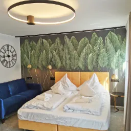 Nowoczesna sypialnia w apartamencie: łóżko z żółtym zagłówkiem, biała pościel, niebieska sofa, zegar ścienny i tapeta z motywem liści bananowca.