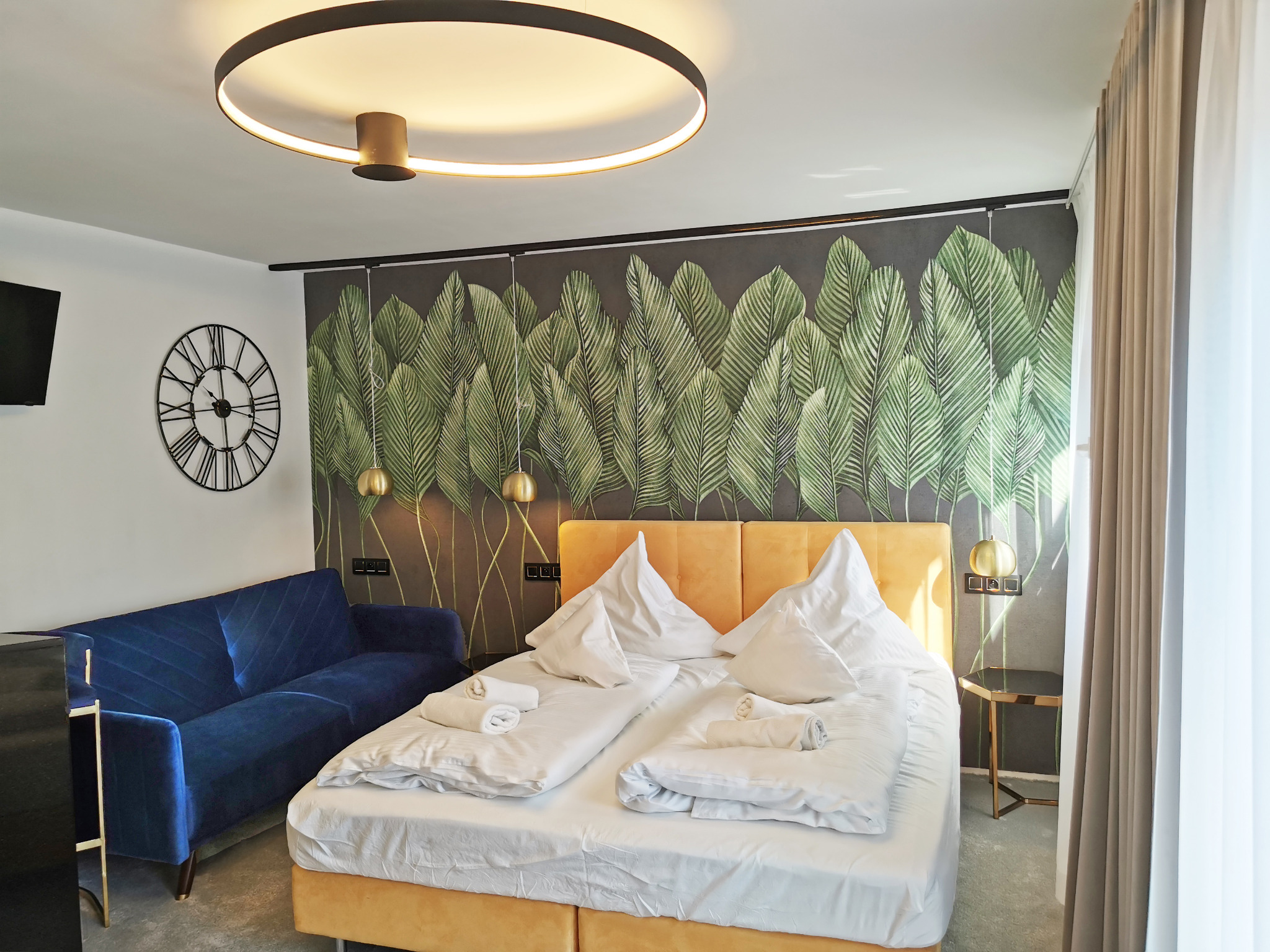 Nowoczesna sypialnia w apartamencie: łóżko z żółtym zagłówkiem, biała pościel, niebieska sofa, zegar ścienny i tapeta z motywem liści bananowca.