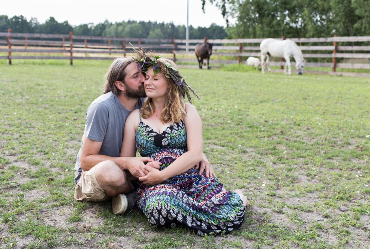 Przyszła mama w kwiecistej sukience i wianku na głowie, siedzi na trawie, całowana w policzek przez partnera, konie i ogrodzenie w tle.