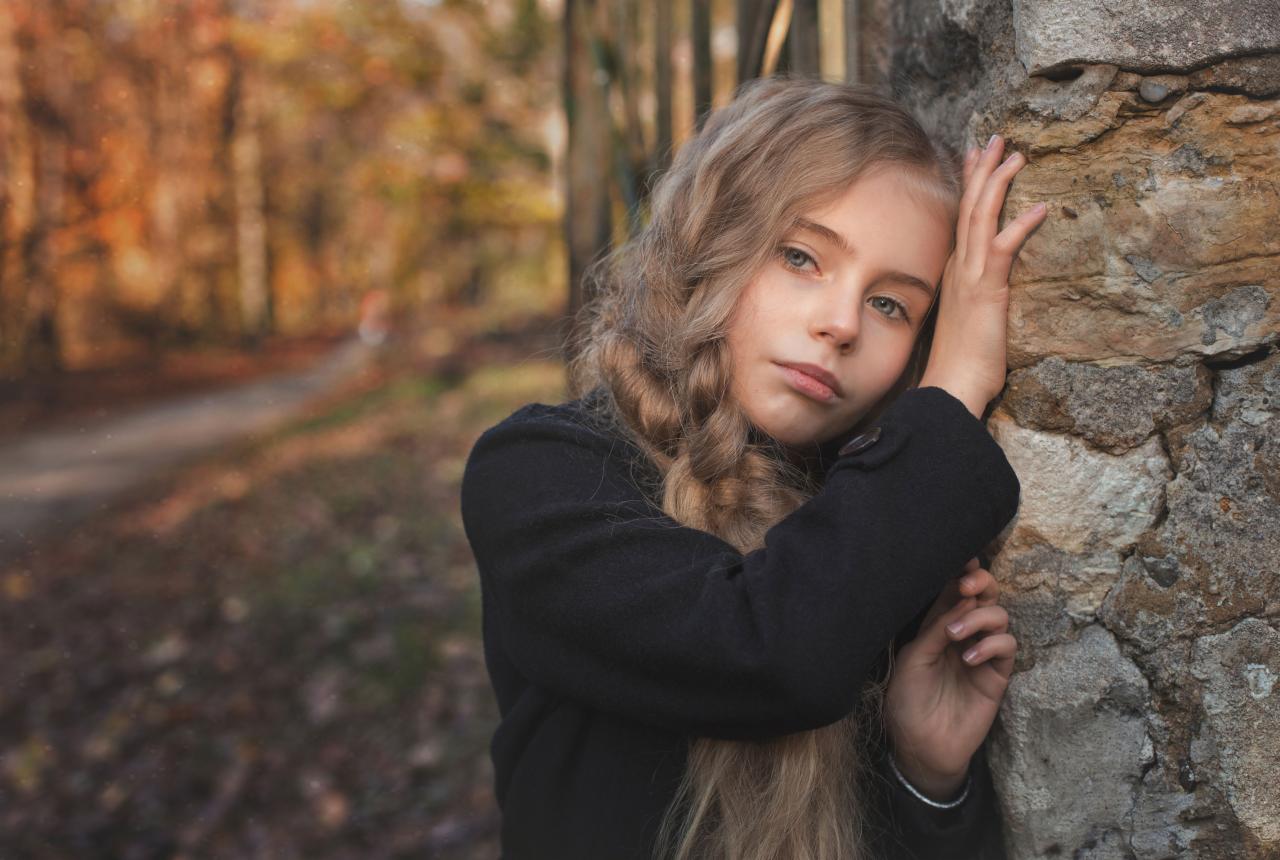 Portret młodej dziewczyny o długich blond włosach, opierającej się o kamienny mur na tle jesiennego lasu, w czarnym płaszczu, z melancholijnym wyrazem twarzy.