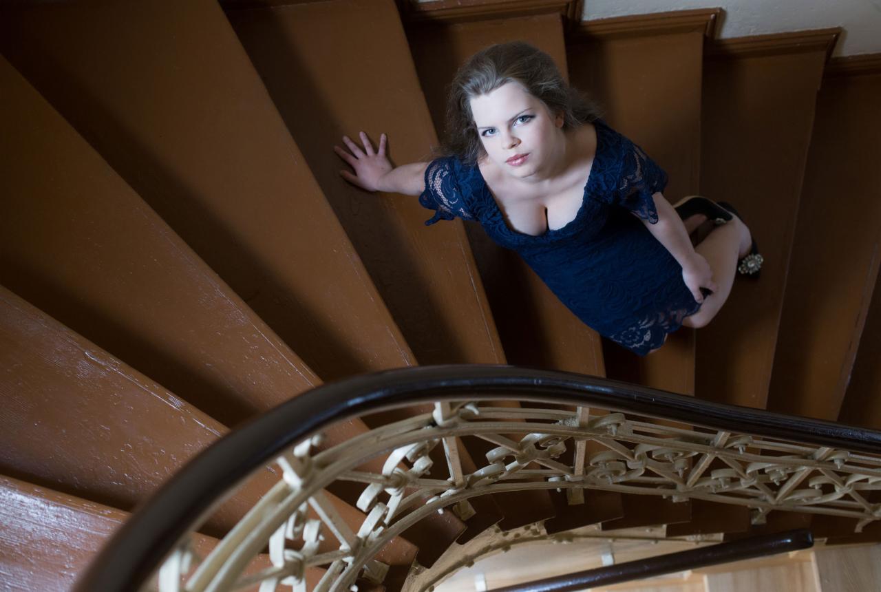 Kobieta w granatowej, koronkowej sukience pozuje na brązowych, spiralnych schodach z ozdobną, białą balustradą, ujęcie z góry.