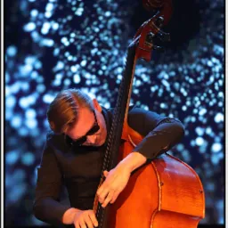 Mężczyzna w czarnej koszuli i okularach gra na kontrabasie na tle rozmytych świateł scenicznych.