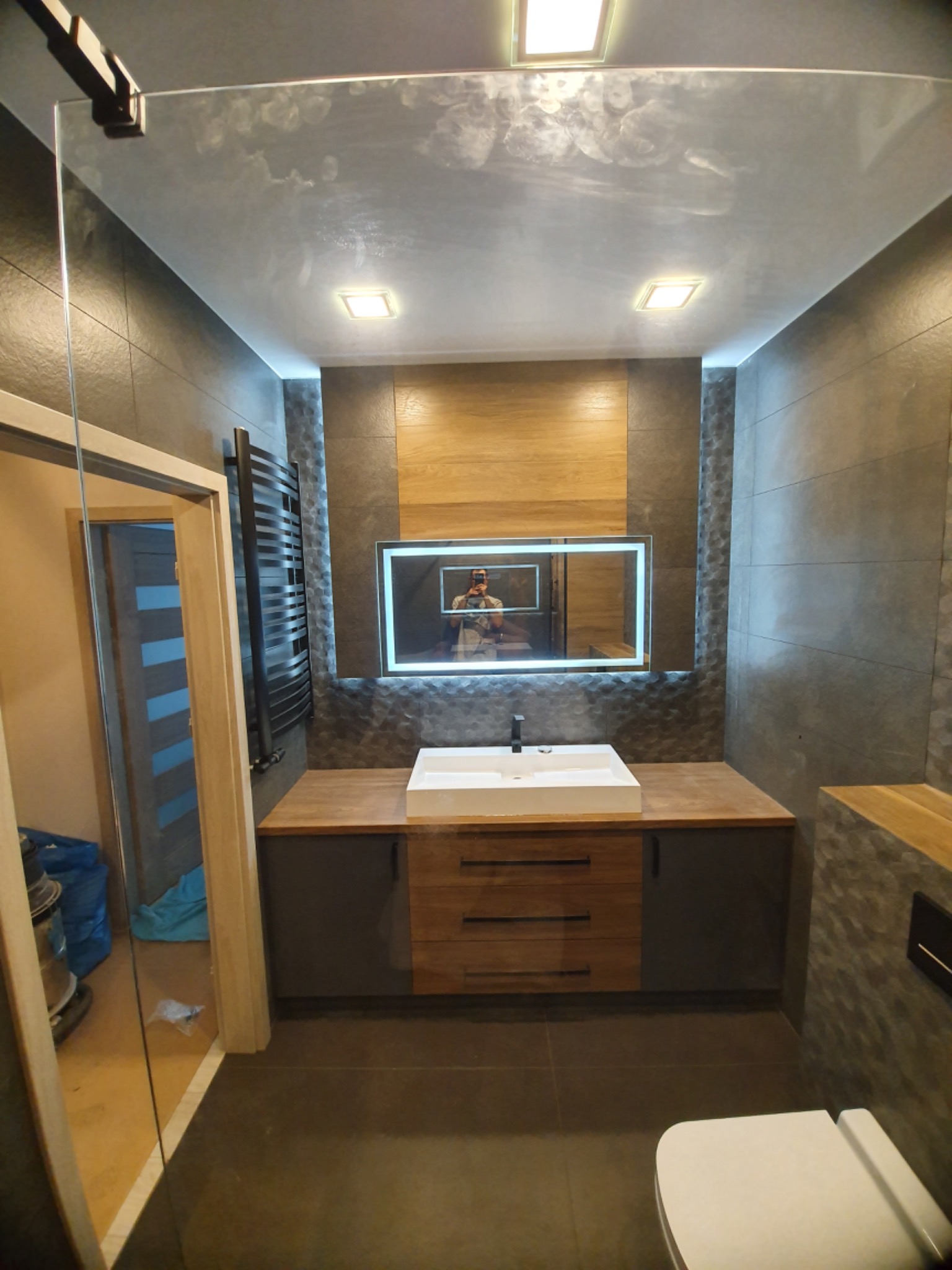 Nowoczesna łazienka z szarą glazurą, drewnianymi akcentami, podświetlanym lustrem i kabiną prysznicową ze śladami po palcach na szkle. Widoczny grzejnik drabinkowy w kolorze czarnym i wisząca toaleta.