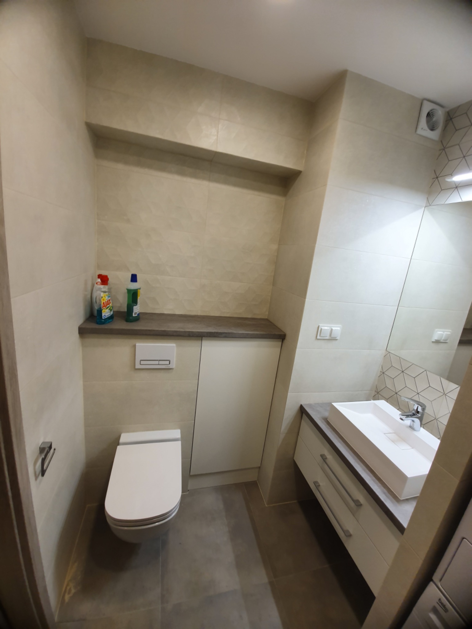 Nowoczesna łazienka z beżowymi płytkami o geometrycznym wzorze, podwieszana toaleta z zabudową, biała umywalka nablatowa z szufladami i dużym lustrem.