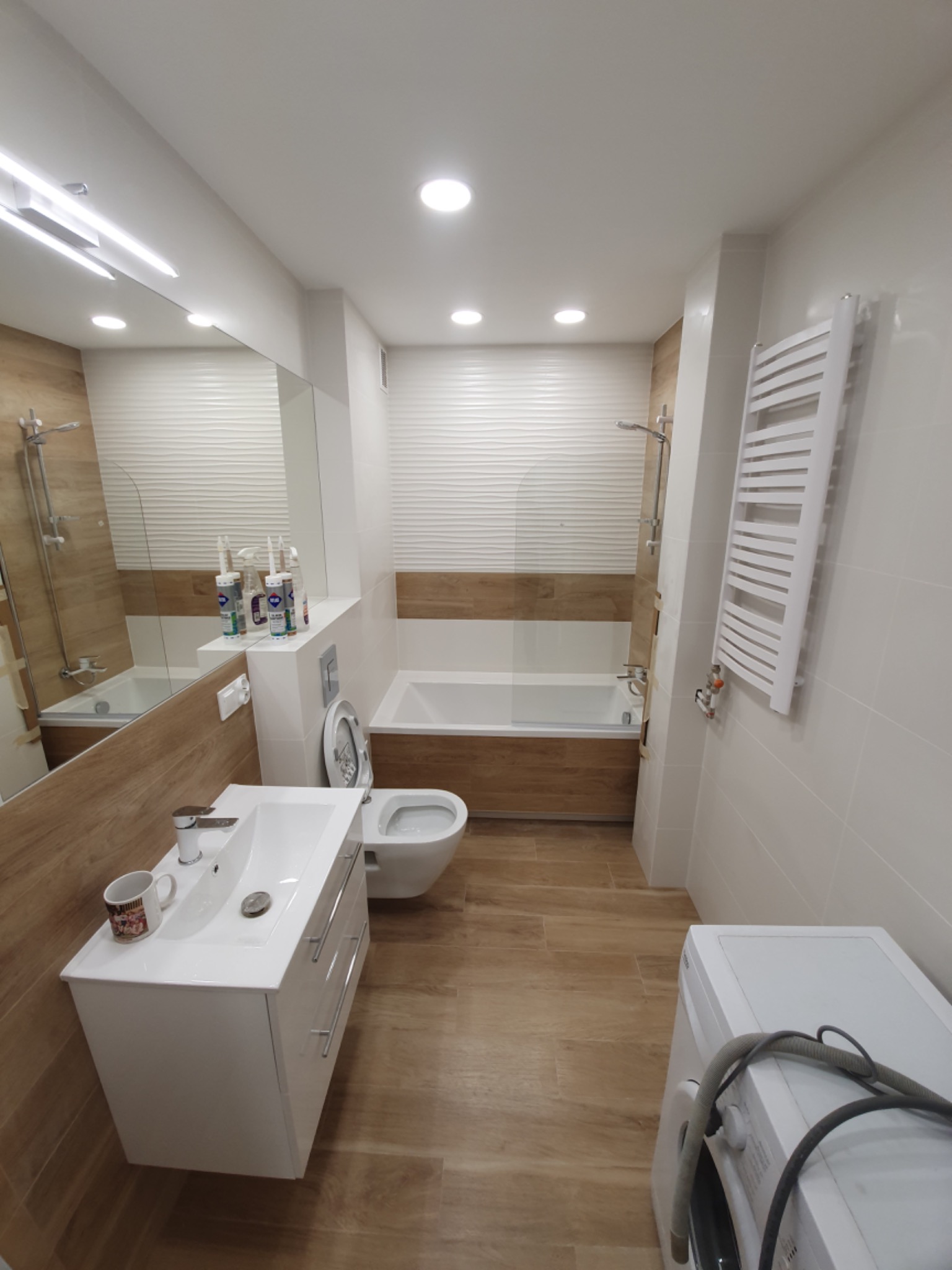 Nowoczesna łazienka po remoncie z wanną, umywalką z szafką, toaletą i pralką. Widoczne białe płytki z drewnopodobnym pasem oraz grzejnik drabinkowy. Na umywalce stoi kubek z nadrukiem.