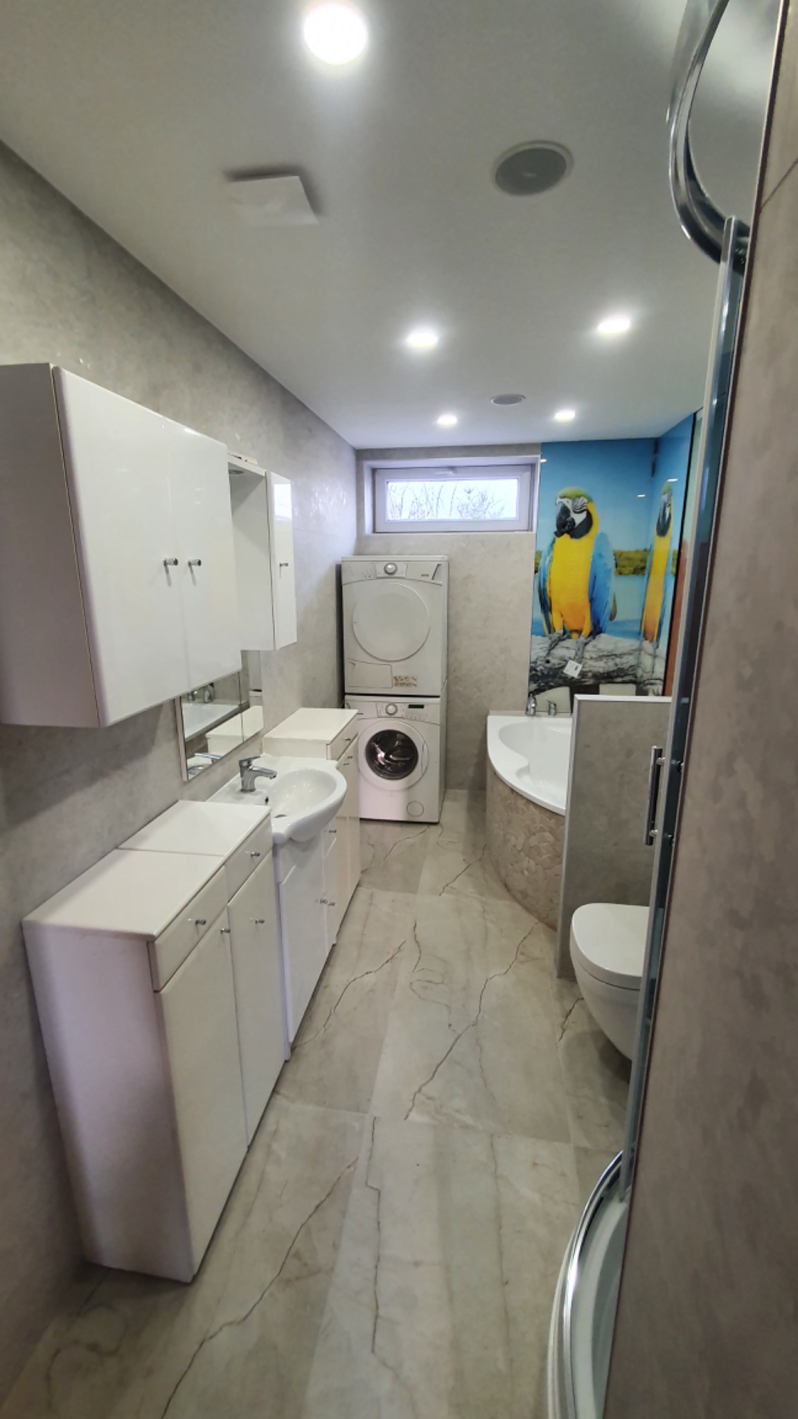 Nowoczesna łazienka z białymi szafkami, umywalką, pralką i suszarką ustawionymi w kolumnie, marmurową podłogą i ścianą z fototapetą z motywem papugi.