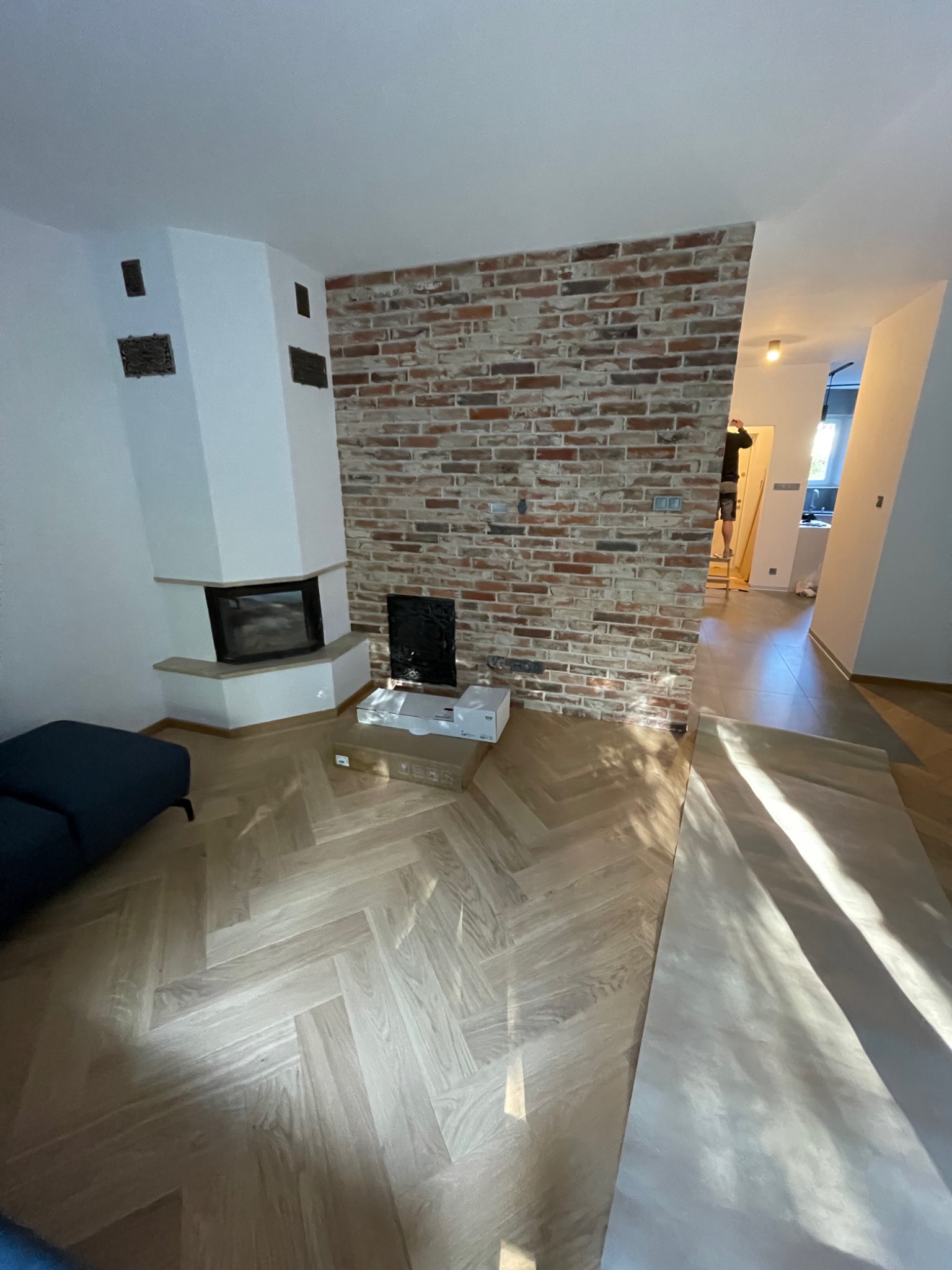Salon po remoncie z nową podłogą ułożoną w jodełkę, ceglaną ścianą i kominkiem, w tle widoczna osoba na drabinie.