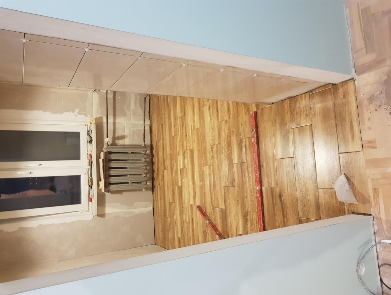 Układanie paneli ściennych imitujących drewno w pomieszczeniu w trakcie remontu, widoczny grzejnik, okno i poziomica.