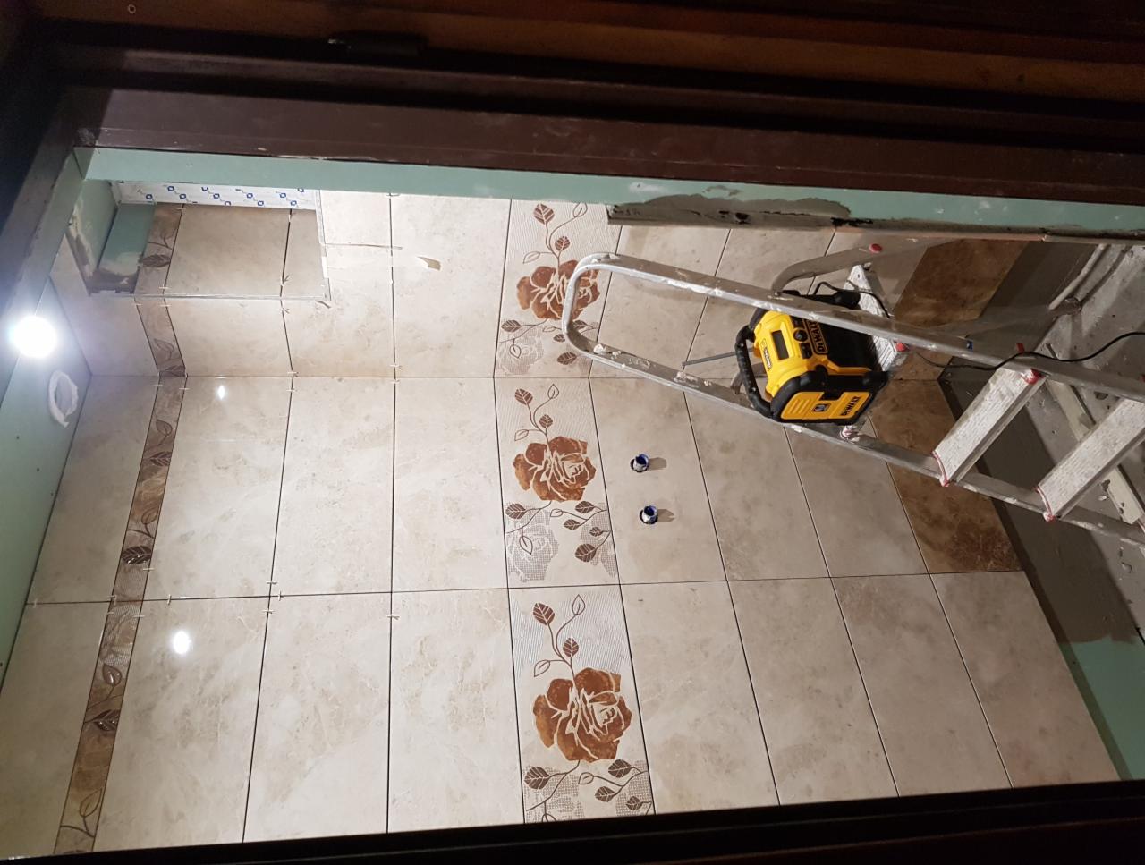 Układanie beżowych płytek z motywem kwiatowym w łazience, widoczna drabina aluminiowa i lampa budowlana DeWalt, fragment surowego sufitu.