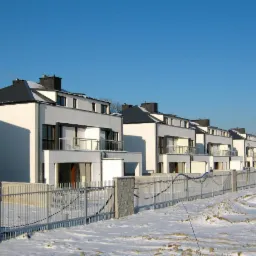 Szereg nowoczesnych, dwukondygnacyjnych domów murowanych z balkonami, ogrodzonych metalowym płotem, w zimowej scenerii z żurawiem budowlanym w tle.