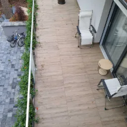 Zadaszony balkon z drewnopodobną podłogą, wyposażony w dwa leżaki, stolik kawowy i grzejnik tarasowy, z widokiem na podjazd dla rowerów i roślinność.