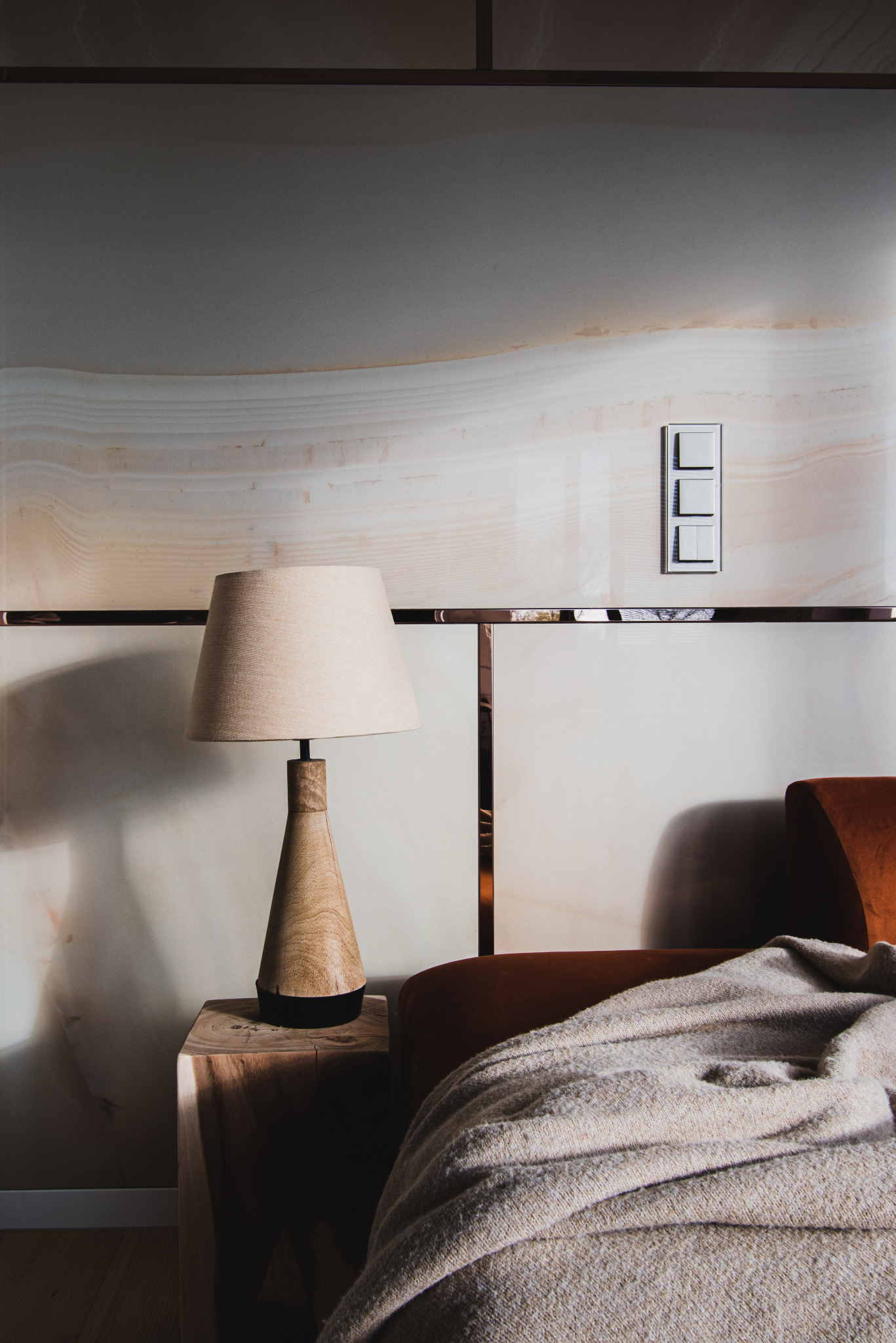Drewniana lampka nocna z beżowym abażurem na surowym, drewnianym stoliku nocnym, obok fragmentu sofy z brązowym obiciem i beżowym kocem, na tle ściany z marmurowym wzorem i trzema włącznikami światła.