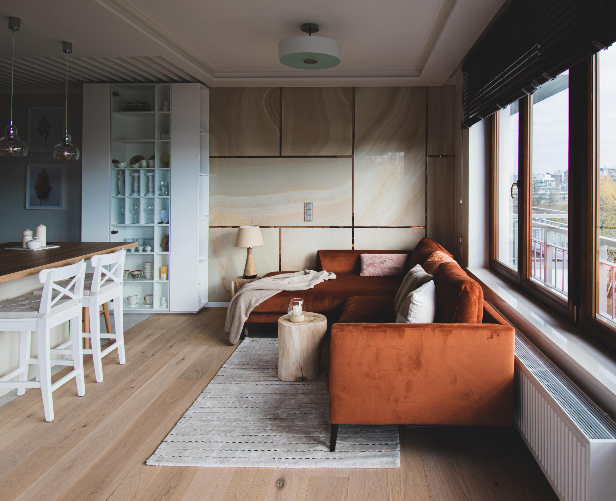 Nowoczesny salon z pomarańczową sofą, drewnianym stolikiem kawowym, marmurową ścianą akcentującą i otwartą kuchnią z białymi krzesłami barowymi.