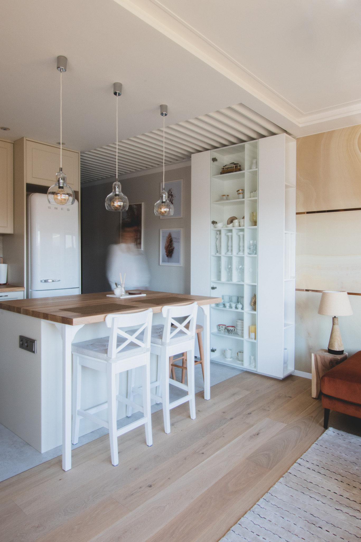Jasna kuchnia z drewnianym blatem, białymi hokerami i lodówką SMEG. Nad blatem wiszą trzy szklane lampy. W tle biała zabudowa z otwartymi półkami, na których stoją szklanki i dekoracje. Na ścianie...